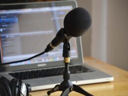 A microphone on a stand with foam windscreen in front of a laptop and headphones.