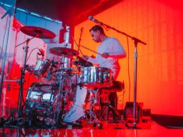 A male drummer performs on a drum kit illuminated by red and blue stage lighting.
