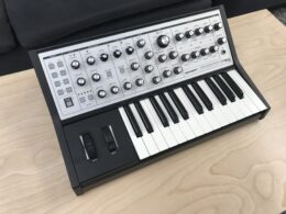 Moog synthesizer with black casing, white keys, and control knobs on wooden surface.