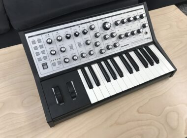 Moog synthesizer with black casing, white keys, and control knobs on wooden surface.