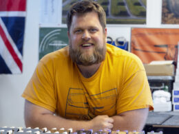 A bearded man in a yellow shirt smiles at a guitar pedal workbench surrounded by electronic equipment.