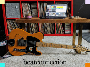 A yellow Squier Telecaster guitar leans against a wooden desk holding a laptop running music software above a red vinyl shelf.