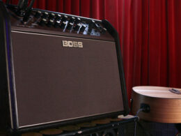 A BOSS acoustic amplifier sits on stage beside a wooden acoustic guitar against a red velvet curtain.