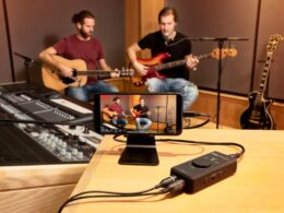 A smartphone on a stand records two musicians performing with guitars through an iRig audio interface in a professional studio.
