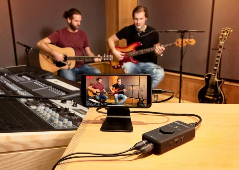 A smartphone on a stand records two musicians performing with guitars through an iRig audio interface in a professional studio.