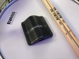 A black boundary microphone sits on a white surface beside wooden drumsticks.