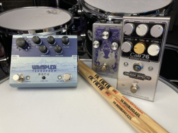 Guitar effect pedals displayed alongside a drum kit and drumsticks on a white surface.
