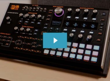Black ASM synthesizer with orange text labels, numerous knobs, buttons, and a grid of colored pads.