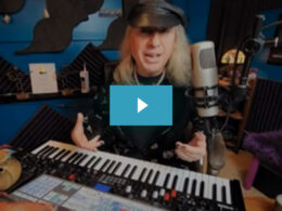 A man with long blonde hair and baseball cap speaks into a microphone at a Yamaha synthesizer in a studio.