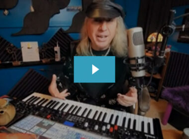 A man with long blonde hair and baseball cap speaks into a microphone at a Yamaha synthesizer in a studio.