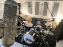 A studio microphone mounted on a shock mount with a blurred drum kit in the background of a recording room.