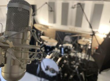 A studio microphone mounted on a shock mount with a blurred drum kit in the background of a recording room.