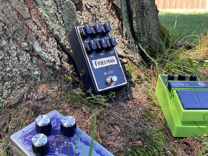 Three guitar effect pedals arranged on the ground outdoors, including a blue Friedman overdrive pedal and two other music gear cases.