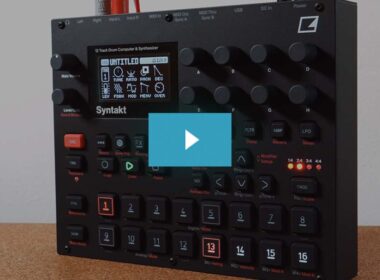 A black Syntakt drum computer and synthesizer with illuminated buttons, knobs, and numbered pads on wood.