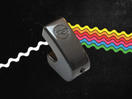 An E-Bow electronic instrument with multicolored wavy lines radiating from it, representing vibrations against a dark background.