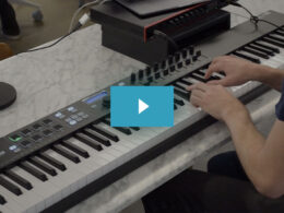A person's hand plays on a Keylab synthesizer keyboard surrounded by music production equipment on a marble desk.