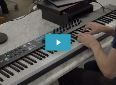 A person's hand plays on a Keylab synthesizer keyboard surrounded by music production equipment on a marble desk.