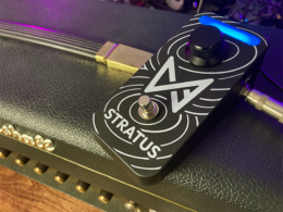 A black Marshall Stratus guitar pedal with gold connectors sits on an olive-green surface under purple stage lighting.