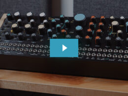 A Taiga synthesizer features cream, blue, and orange knobs on a wooden base.