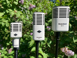Three vintage ribbon microphones on stands are surrounded by green foliage and pink flowers.