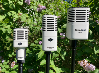 Three vintage ribbon microphones on stands are surrounded by green foliage and pink flowers.