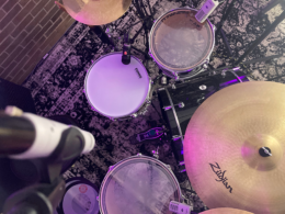 An overhead photograph of a drum kit bathed in purple and pink lighting.