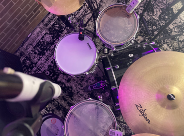 An overhead photograph of a drum kit bathed in purple and pink lighting.