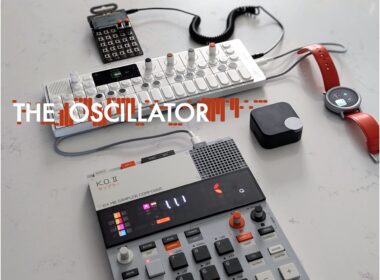Electronic music production equipment including a white keyboard synthesizer, black sampler, MIDI grid controller, mouse, and smartwatch.