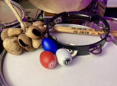 Percussion instruments including maracas, a tambourine, Vic Firth drumsticks, and colorful balls arranged on a white surface.