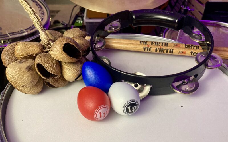 Percussion instruments including maracas, a tambourine, Vic Firth drumsticks, and colorful balls arranged on a white surface.