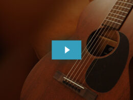 An acoustic guitar with warm brown tones is displayed at an angle with a blue play button overlay in the center.