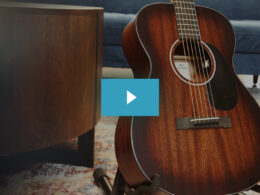 A reddish-brown acoustic guitar sits on a patterned rug in a home interior with wooden furniture and blue upholstery.