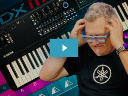 Man wearing blue glasses poses beside a Yamaha MODX-M synthesizer keyboard on a dark background.
