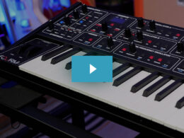 A black music synthesizer with illuminated controls sits behind a white keyboard in a studio with blue lighting.