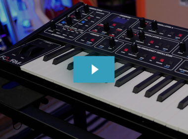 A black music synthesizer with illuminated controls sits behind a white keyboard in a studio with blue lighting.