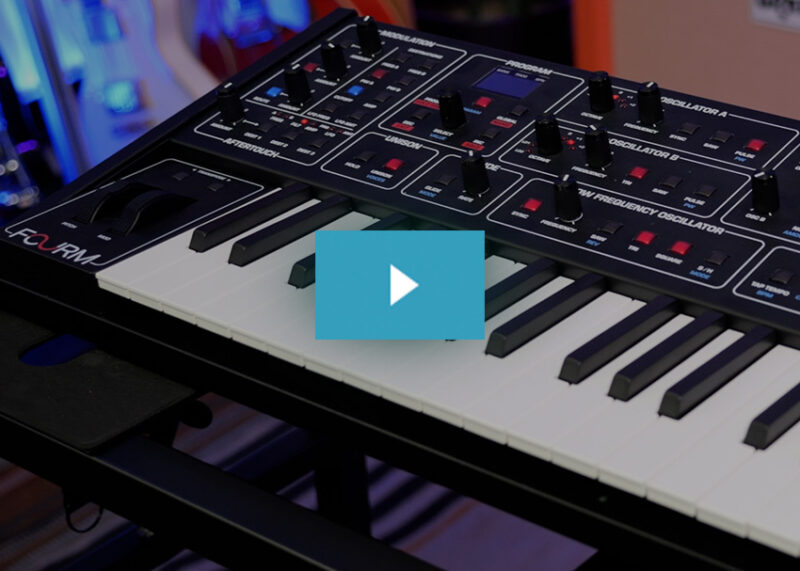 A black music synthesizer with illuminated controls sits behind a white keyboard in a studio with blue lighting.