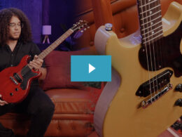 A musician with curly hair and glasses plays a red electric guitar on a red leather couch surrounded by guitars.