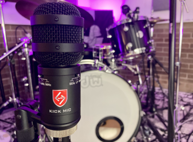 A blue recording microphone stands in front of a drum kit in a studio.