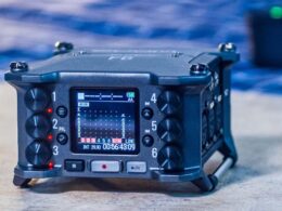 A blue portable digital audio recorder displays timecode and audio levels on its LCD screen among multiple control knobs and buttons.