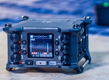 A blue portable digital audio recorder displays timecode and audio levels on its LCD screen among multiple control knobs and buttons.