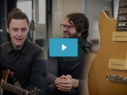 Two men smile together in a music studio surrounded by gold electric guitars.