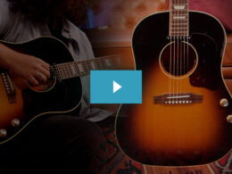 A musician plays a black acoustic guitar next to a larger sunburst acoustic guitar in a warm studio environment.