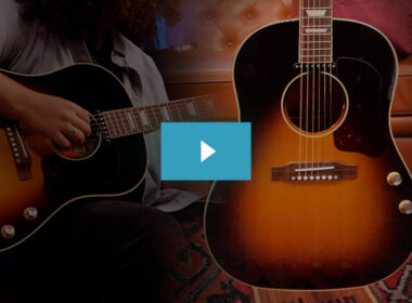 A musician plays a black acoustic guitar next to a larger sunburst acoustic guitar in a warm studio environment.