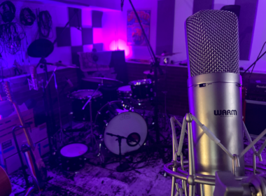 A large professional microphone stands in a dark recording studio bathed in purple and blue neon.